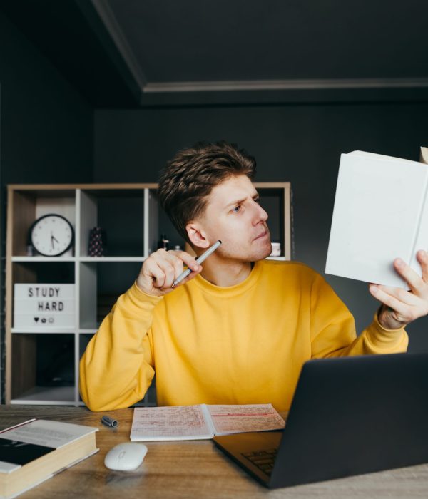 Smart young student is sitting at home at a table with a laptop and books on background of a cozy bedroom,reading a book with a focused face and holding a pen.Pensive guy studies at home in quarantine